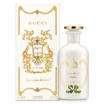 A white Gucci perfume bottle with gold accents stands next to its matching ornate box. The label reads "Love at your Darkest." Both feature decorative gold detailing and branding.