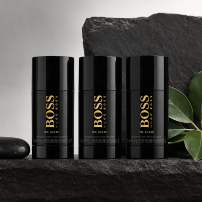 Three black cylindrical deodorant sticks labeled "BOSS The Scent" stand on a dark stone surface with green leaves in the background.