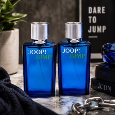 Two bottles of Joop Jump EDT for Men, 30ml travel size, are displayed side by side on a marble surface with books, a towel, a plant, and a sign in the background.