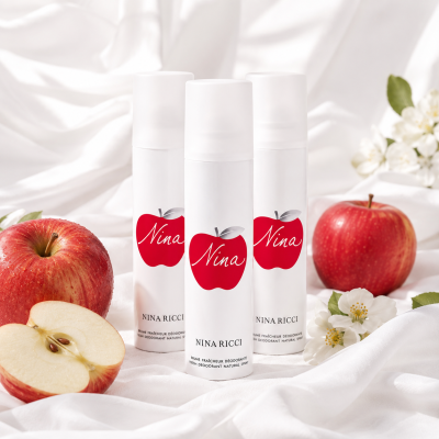 Three bottles of Nina Ricci Nina Deodorant Spray for Her 150ml are displayed among apples and apple blossoms on a white fabric background.
