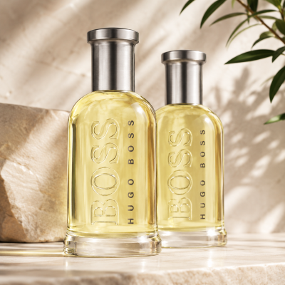 Two bottles of Hugo Boss Bottled EDT for Men (100ml each), featuring clear glass and silver caps, are displayed side by side on a stone surface with a plant in the background.
