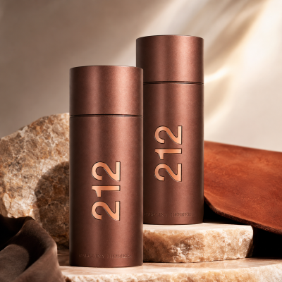 Two cylindrical brown bottles of Carolina Herrera 212 Sexy Men Eau de Toilette (100ml spray) stand on stone slabs, with “Carolina Herrera” etched on the base. Draped brown and beige fabric appears in the background.