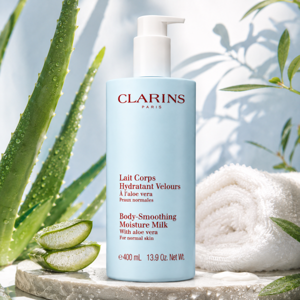 Three bottles of Clarins Body Smoothing Moisture Milk With Aloe Vera (400ml each) are arranged on a stone surface, surrounded by aloe leaves and a rolled white towel, illuminated by natural light.