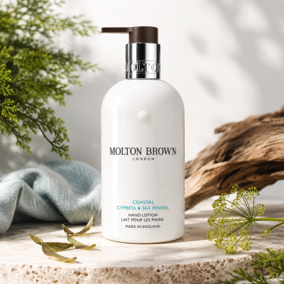 Two bottles of Molton Brown Coastal Cypress & Sea Fennel Bath and Shower Gel (300ml, Unisex) rest on a surface with green plants, a light blue cloth, and driftwood in the background.