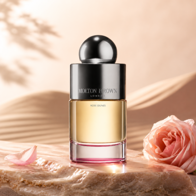 A bottle of Molton Brown Rose Dunes EDT 100ml, Unisex placed on a marble surface beside a pink rose and petal, with a softly lit background.