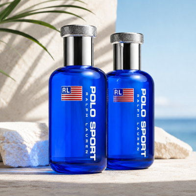 Two bottles of Ralph Lauren Polo Sport 75ml EDT Spray for Men are displayed on white stones, with a blurred ocean and sky background and a palm leaf in the upper left corner.