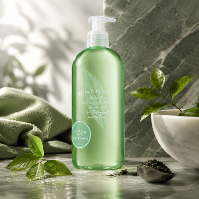 Three green bottles of Elizabeth Arden Green Tea Bath & Shower Gel for Her, 500ml each, are arranged on a marble surface alongside tea leaves, a towel, and a bowl of loose green tea leaves.