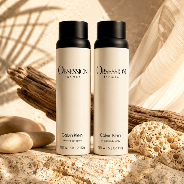 Two bottles of Calvin Klein Obsession for Men body spray stand on sand beside rocks, driftwood, and coral, with soft sunlight illuminating the scene.