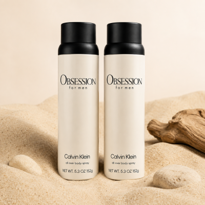Two bottles of Calvin Klein Obsession for Men body spray stand upright on sand next to a piece of driftwood.