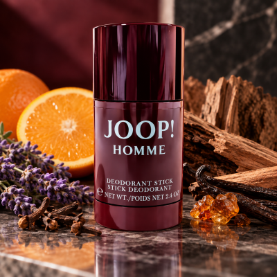 A JOOP! Homme deodorant stick sits on a marble surface with orange slices, lavender, amber resin, and wood in the background.