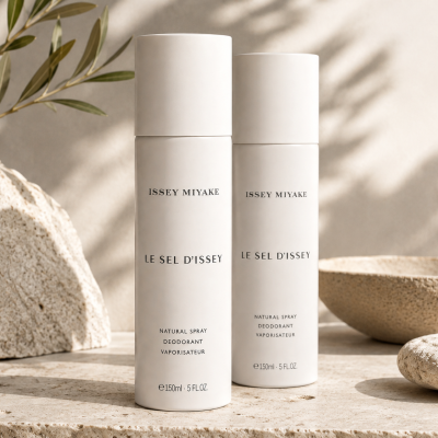 Three bottles of Issey Miyake Le Sel D’Issey Deodorant Spray for Men, 150ml, stand on a beige surface, accompanied by textured stone decor and bathed in soft sunlight.