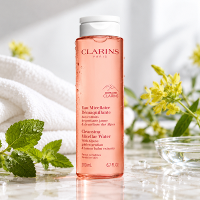 A bottle of 3x Clarins Cleansing Micellar Water 200ml on a marble surface with pink liquid, mint leaves, a white towel, and yellow flowers.